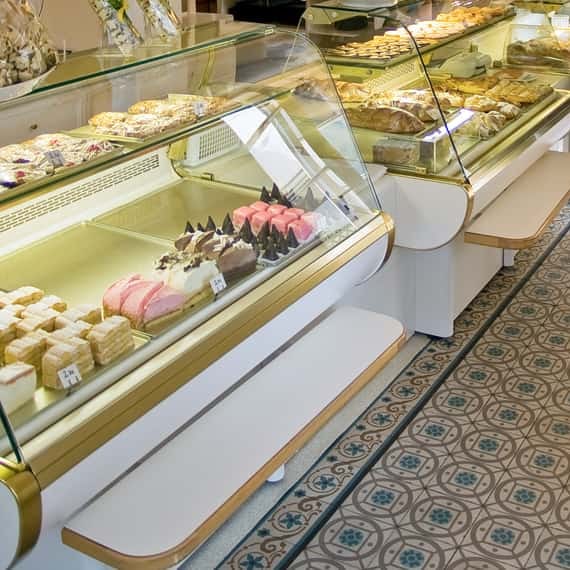 Blick in die Vitrine: Cremeschnitten, Indianer und rosa Mehlspeisen über dem historischen Fliesenboden