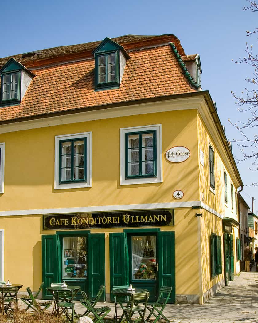 Das gelbe Eckhaus der Café-Konditorei Ullmann in Baden mit dunkelgrünen Fensterläden und Schanigarten