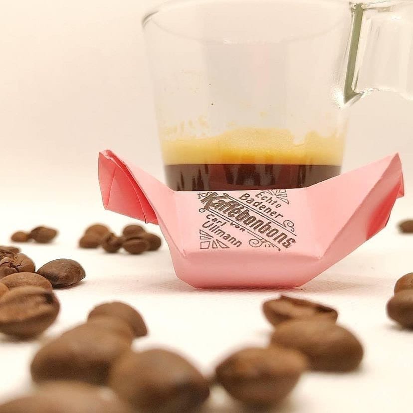 Ein Badener Kaffeebonbon im rosa Wickelpapier mit dem Aufdruck „Echte Badener Kaffeebonbons von Carl Ullmann“ vor einer Espressotasse